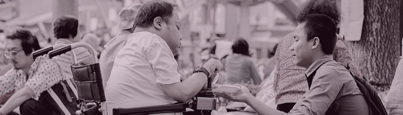 Image of two men sitting in among a crowd of people in an outdoor space. One man is helping to hold a plate for the other who is in a wheelchair with visible mobility barriers